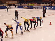 Cinq patineurs alignés sur la ligne de départ, avec deux juges derrière et sur le côté.