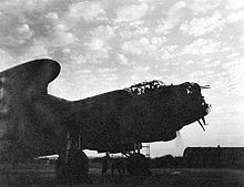 Front three-quarter view of military aeroplane with propellers spinning, silhouetted against the sky