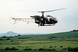 Hélicoptère de couleur verte avec l'inscription « Bundesgrenzschutz » sur le flanc vole le long d'une clôture grillagée derrière laquelle se trouvent deux soldats est-allemands et un camion.