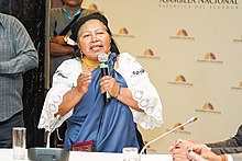 A woman in national costume speaks into a microphone in front of a backdrop bearing the words "Asamblea Nacional Repulica del Ecuador"l