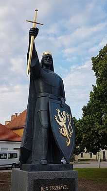 Fotografie černé sochy muže stojící na podstavci. Muž v levé zvednuté ruce drží zlatý meč a v pravé ruce štít s zlatým reliéfem znaku Českého království. Muž má plášť, dlouhé vlasy a na hlavě zlatou korunu.