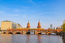 Frontale Farbfotografie von einer roten Backsteinbrücke mit einer gelben U-Bahn in der Mitte. Die Backsteinbrücke hat zwei große Türme und eine Unterführung für Autos und Fußgänger.