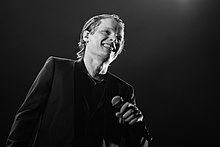 Photographie en noir et blanc de Bénabar debout, sur scène, et souriant.