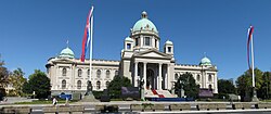 Duży biały budynek z zielonymi wieżami i kopułami, przy wejściu kolumnada, po prawej i po lewej stronie budynku flagi narodowe na maszcie.