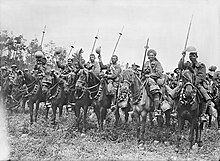 Indian cavalrymen sit on their horses in a line, holding lances and waving their helmets