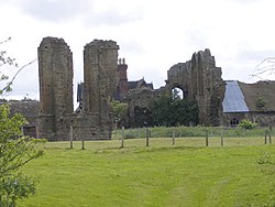 A photograph showing two tall sections of rough stonework, one housing a tall window without top, the other two gothis arches, behind a wire fence.