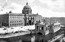 Schwarzweißfotografie von einem dreigeschossigen hohen Gebäude. Über dem Eingang, der wie ein griechischer Triumphbogen aussieht, befindet sich ein weiteres Geschoss mit einer Kuppel. Vor dem Gebäude sind eine breite Straße und ein bebautes Flussufer mit einem Kolossalportal mit Reitern und weiteren Figuren.