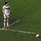 Photographie d'un joueur qui se concentre, le regard vers les poteaux de but, avant de prendre l'élan pour tenter un but de pénalité en frappant le ballon posé en travers sur un tee.