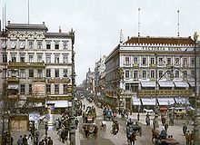 Farbfotografie mit Blick auf eine Kreuzung mit vielen Kutschen und Passanten. Im Vordergrund ist an der rechten Fassade die Aufschrift „Victoria Hotel“ und an weiteren Häuserfassaden sind Reklametafeln.