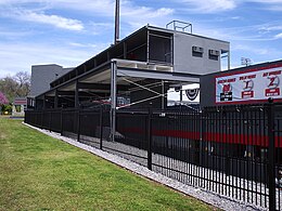 The back side of a freshly painted dark gray metal and concrete structure with red and white accents. Concession stands can be seen on the first level, seating on the second, and sky boxes on the third.