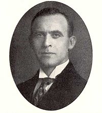 A black and white photograph of a dark haired man in a dark business suit with a white shirt and a tie underneath.