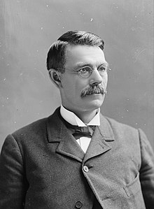 Head of a 40sh man with gray hair, squinting through eyeglasses