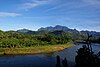 A river cuts through a forest that blankets the base of a mountain range, with a massive mountain range in the background.