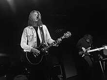 Long haired male singer with Les Paul guitar standing on stage at the microphone in a black and white shot taken from a lower position in the audience