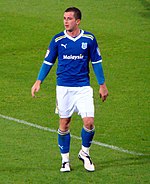 A young man wearing blue and white shirt, shorts and socks, standing on a grass field