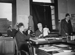 several men seated at tables in a room with Kippenberger standing on right