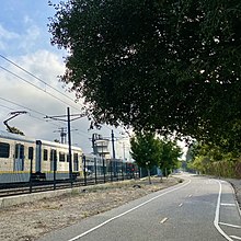 E Line train passes Expo Bike Path in Culver City; Hayden Tract architectural landmark visible in distance, native oak tree overhangs the path