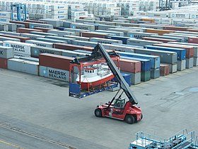 Reach stacker carrying a small vessel on a flat rack, in a container yard