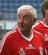 The head and upper torso of a gentleman in his late 50s. He has short white hair and is wearing a red football shirt, which has the 利物浦足球俱乐部 crest on the left breast, and a crest on the right breast that says "利物浦 Legends". A white logo of the Adidas sponsor is visible in the centre of the shirt, and three white stripes are present on the shoulder.