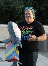 Photographie d'une femme aynt des cheveux longs attachés en chignon dont une partie sont teints en bleus, un tshirt noir avec "Stand against hate" écrit en arc-en-ciel et un jean. Elle tient contre elle un requin en peluche et tient dans la main un sac en papier arc-en-ciel