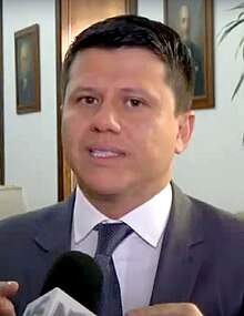 A man in his forties stands in front of a microphone wearing a business suit. He had tan skin and short black hair. There are portrait paintings visible in the background.