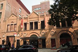 The right half of an old, sturdy-looking 4-story, 5-bay building, faced with partly soiled beige brick and architectural decorations. The right bay has a gabled fifth floor with 3 small windows. A fire escape runs down the front. At street level 1½ large doorways are visible. One of the 20 or so windows is boarded, another is painted black.