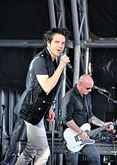 A dark-haired man singing into a microphone and a bald man playing a guitar