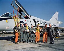 The astronauts pose in front of a delta-winged light blue-gray jet aircraft, holding their flight helmets under their arms. The three Navy aviators wear orange flight suits; the Air Force and Marine ones are green.