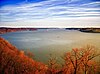 View of a very wide river lined with bare trees in reddish light under a blue sky streaked with clouds