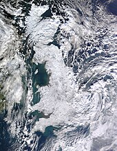 A satellite photo of Great Britain and Ireland. Great Britain is white as a result of snow cover and Ireland is mostly green with some snow cover in the east. Cloud is scattered across the surrounding sea and eastern Ireland, as well as the east coast of Great Britain (especially the southeast).