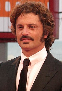 photo en buste d'un homme jeune, cheveux frisés châtains et moustache ; veste et cravate noires avec chemise blanche