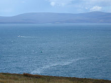 The attack site today, seen from a cliff above the bay. A small green wreck buoy is a few hundred metres away. A thin slick of oil is on the surface of the sea.