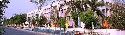 A campus building right beside the main road, surrounded by trees and flags polled along the footpath