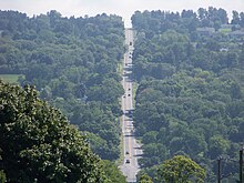 A two-lane highway descends sharply into a valley, where it intersects another roadway at the base of the valley. From there, it ascends the opposite side of the valley and disappears into the background.