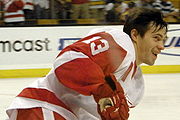 A side shot of an ice hockey player. He has medium length brown hair and is wearing a red and white uniform.