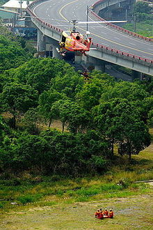 A UH-1 helicopter in midair hanging a rescue officer, ready to fetch the wounded on the ground.