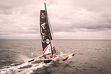 Trimaran vu de l'arrière, filant sous voile vers un ciel nuageux tout rose.