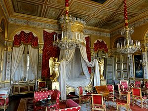 Farbfotografie eines Innenraums mit einem weißen Himmelbett an der hinteren Wand, dessen Vorhänge von goldenen Engeln gehalten werden. An beiden Seiten des Bettes sind zwei Fenster mit weißen Vorhängen und roten Kurzgardinen mit goldenem Muster. Die Wände und Decke sind mit gold-weißen Ornamenten versehen. Von der Decke hängen zwei Kronleuchter und im Vordergrund stehen rote Stühle und Sessel mit goldenem Muster.