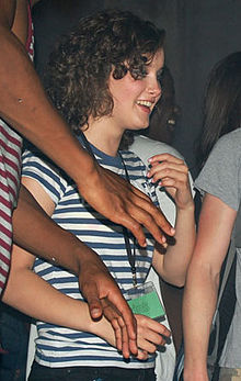 A young, caucasian female with long, dark hair whom looks away from a camera against a backdrop of individuals.