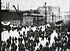 Trabajadores de Harland and Wolff a la salida del astillero, hacia abril de 1911. A la izquierda, se observa el SS Nomadic, transbordador del Olympic y del Titanic, durante su acondicionamiento. Al fondo, se observa el pórtico Arrol, con la proa del Titanic perfectamente visible.