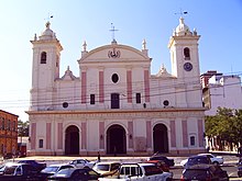 Photo of a Spanish church with two towers, and three arched entrances. In front are parked cars.