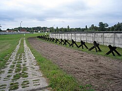 Bande de terre labourée longée par une route bétonnée d'un côté et une rangée d'obstacles anti-véhicules et un mur, de l'autre côté.