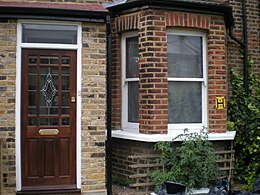 A edificiu de lladriyu marrón. Visible is a door at left, with a bowfront window at right; various tomato plants are also visible growing in front of the window.