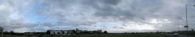 Photo panoramique : vaste ciel tourmenté, en arrière plan, mais en premier plan ligne plutôt sombre d'immeubles entourés d'arbres et une colonne de voitures