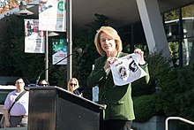 Woman standing in front of a podium holding an upside-down piece of paper which commemorates the addition of Seattle to the National Hockey League.