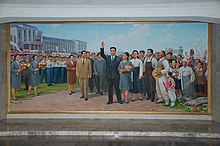 Peinture murale montrant Kim Jong-Il au centre, entouré par une foule compacte de personnes, l'université est visible dans le fond.