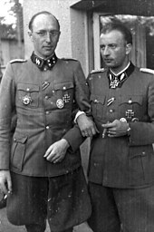 A black-and-white photograph of two men wearing military uniforms with various military decorations attached to their uniforms. The man on the left is wearing glasses and is smoking a cigarette, the man on the right is smoking a cigar.