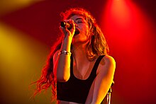 A picture of Lorde performing at a concert. Her face is captured singing in a dramatic pose, wearing a black crop top with yellow and red lights illuminating the stage.