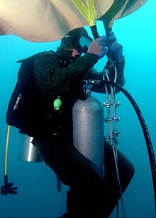 Underwater, a US Naval diver in a scuba suit with mask, oxygen tank, and regulator, is attaching a large, upside-down beige bag to braided metal chain.