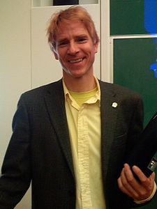 Christof Koch wearing a yellow t-shirt under a yellow shirt, with a dark blazer, holding a laptop computer in his left hand, grinning at camera